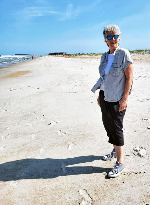 An older woman stands on a sandy beach, posing for the camera.